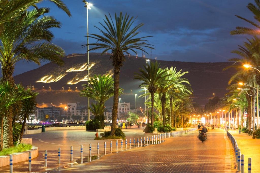 Promenade illuminée d’Agadir la nuit, bordée de palmiers, avec des lumières urbaines chaleureuses et la colline en arrière-plan affichant une inscription lumineuse, créant une ambiance moderne et touristique.
